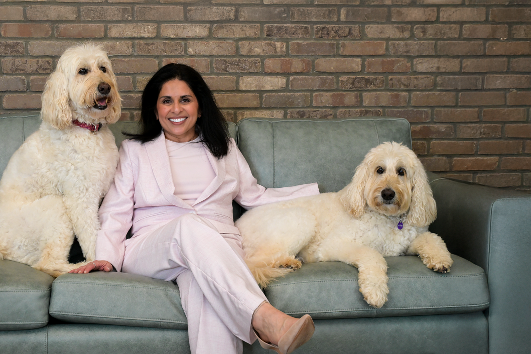 Dr. Veena Bhat sitting on a light teal couch in a pale pink suit, smiling between two large white fluffy dogs, in front of a brick wall – North Fulton Smiles