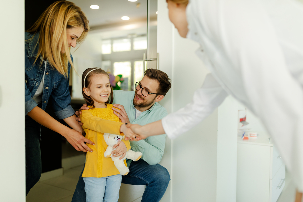 A cheerful young girl holding a stuffed toy is warmly greeted with a handshake by a dental professional, as her smiling parents supportively stand nearby in a modern dental office – Family Dentistry